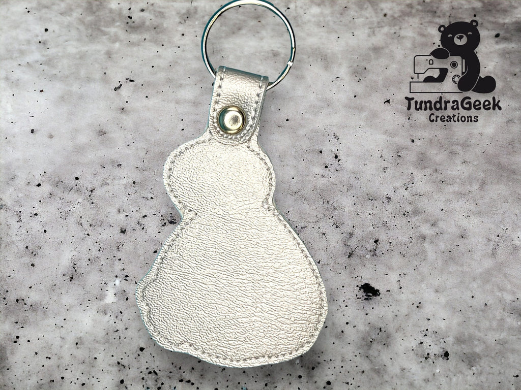 A silver snowman-shaped leather keychain with a snap button, placed on a speckled gray background next to the TundraGeek Creations logo.
