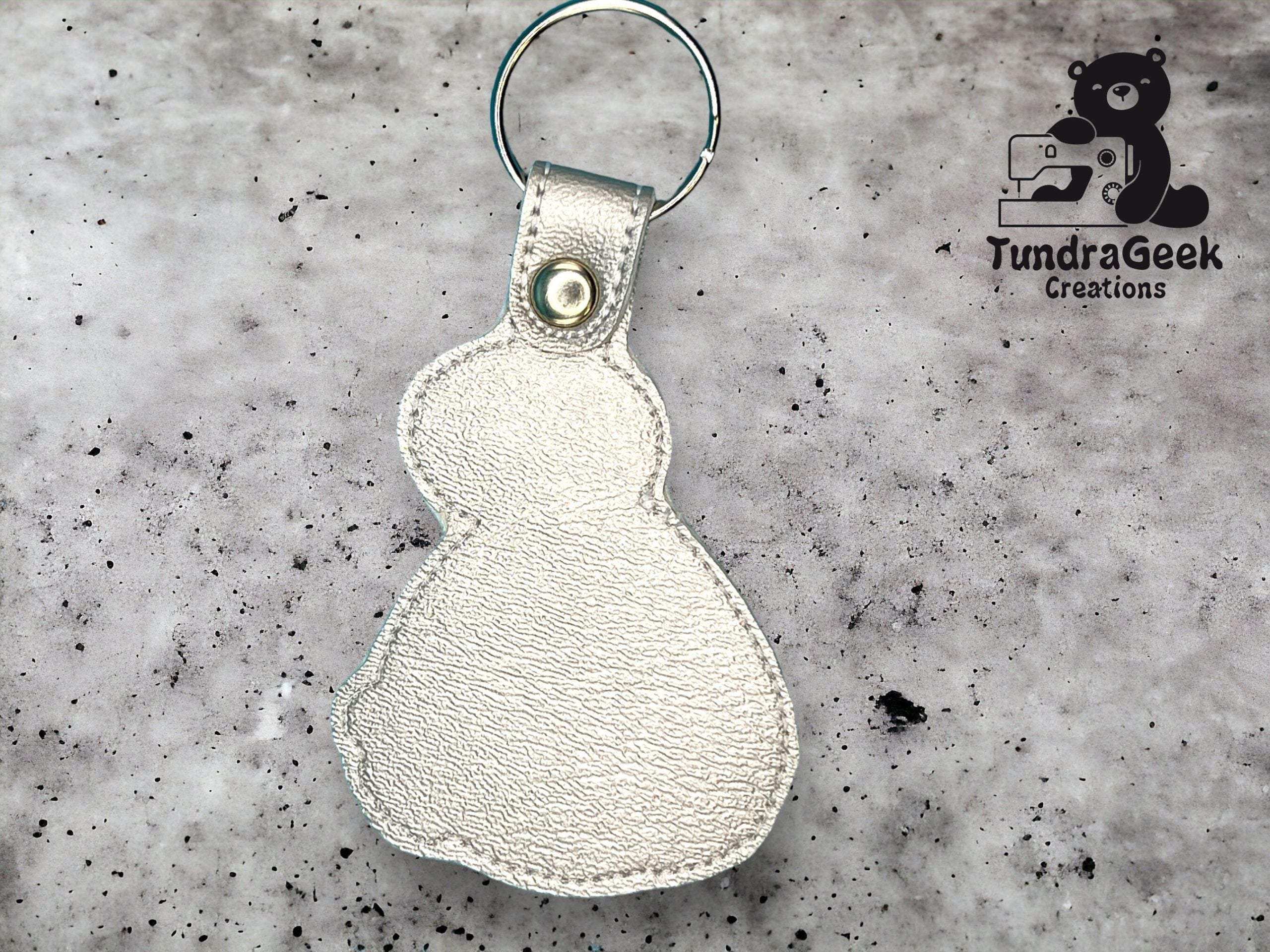 A silver snowman-shaped leather keychain with a snap button, placed on a speckled gray background next to the TundraGeek Creations logo.