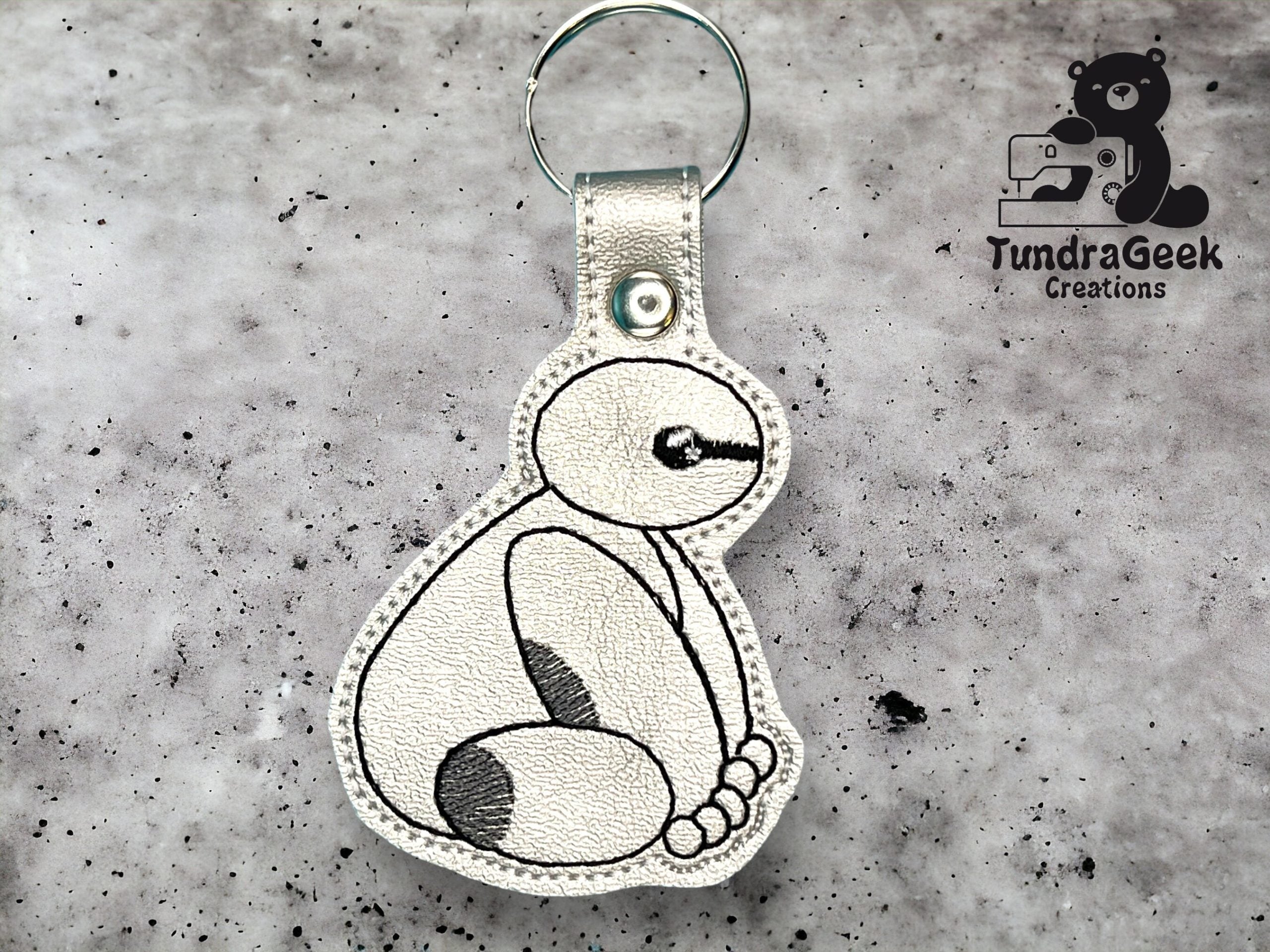 A keychain featuring a stitched outline of a sitting robot character on metallic silver material, displayed on a textured gray background with the TundraGeek Creations logo.