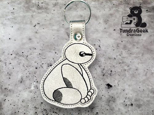 A keychain featuring a stitched outline of a sitting robot character on metallic silver material, displayed on a textured gray background with the TundraGeek Creations logo.