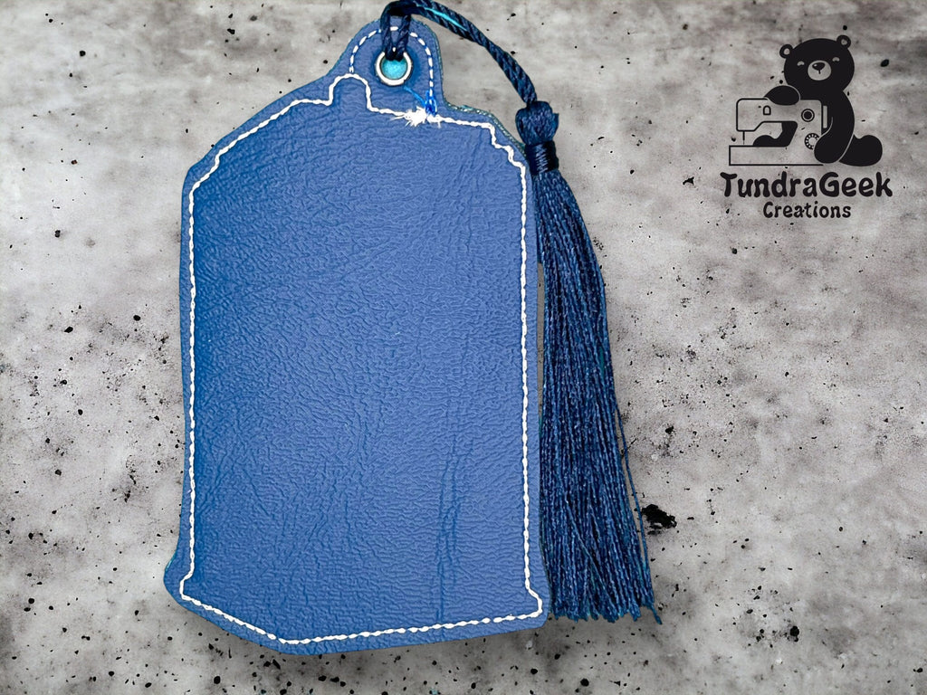Blue leather keychain shaped like Michigan with white stitching and a blue tassel, against a gray background with the TundraGeek Creations logo in the corner.
