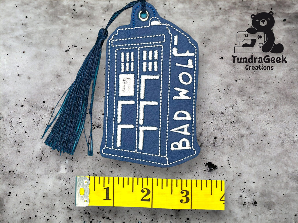 A blue embroidered keychain in the shape of the TARDIS with BAD WOLF text, next to a yellow measuring tape showing a length of about 4 inches, and a TundraGeek Creations logo.