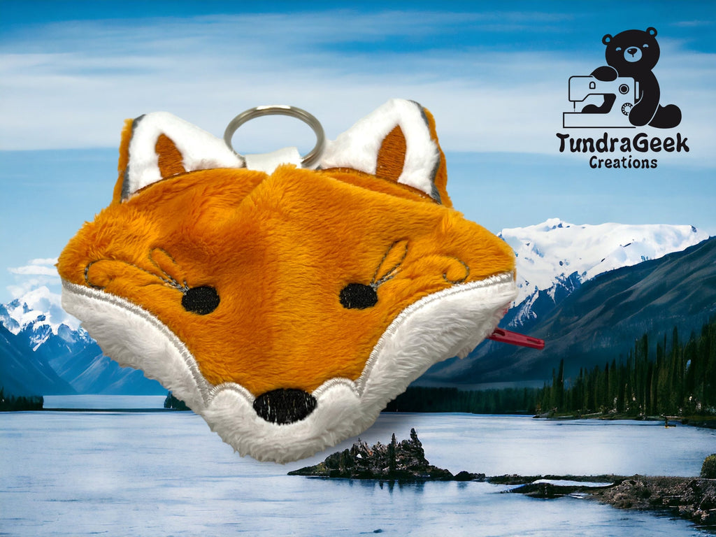 A plush fox-shaped coin pouch with a zipper sits in front of a scenic mountain lake backdrop; TundraGeek Creations logo appears in the top right corner.