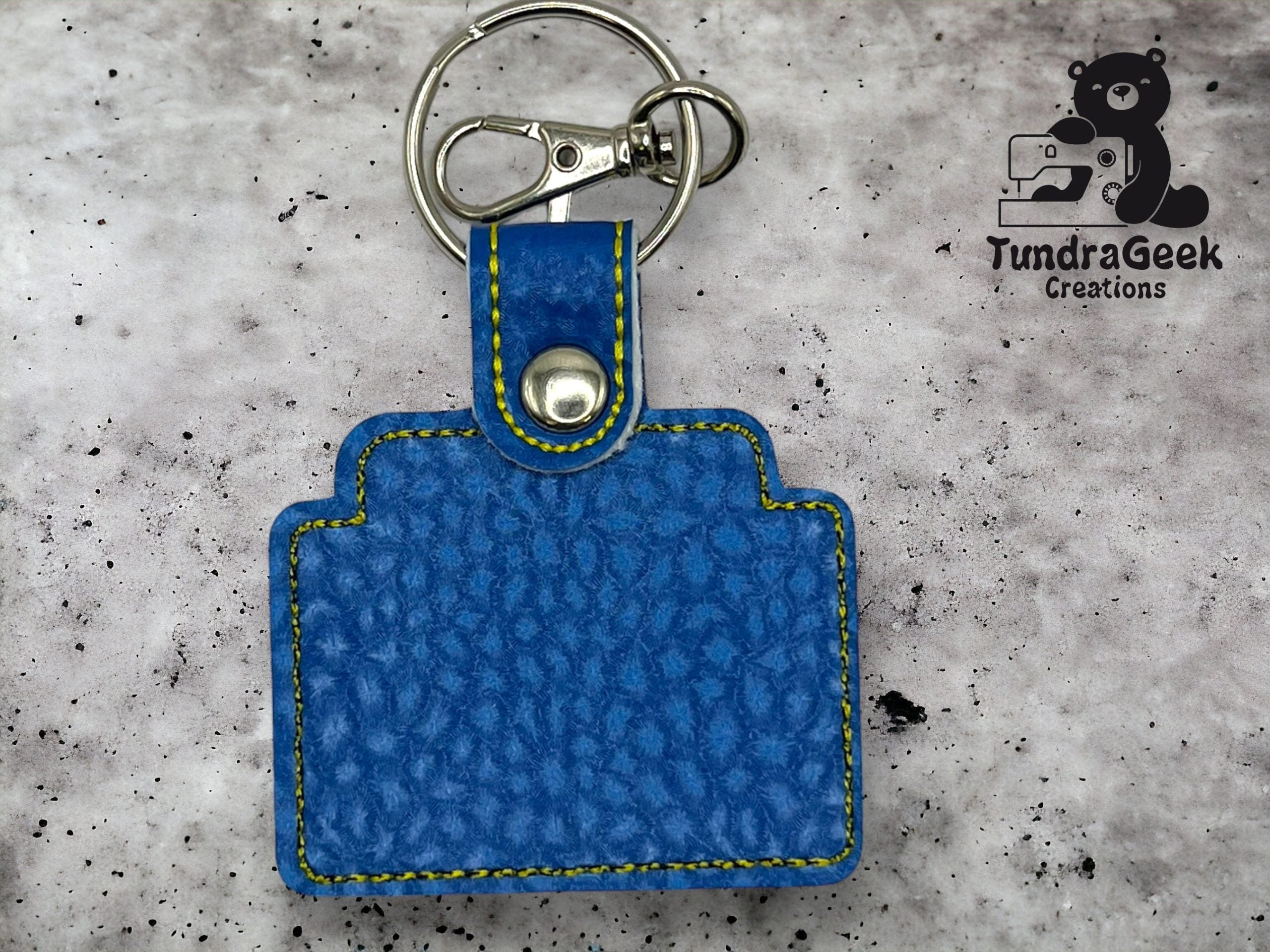 A blue, textured camera-shaped keychain with yellow stitching lies on a gray speckled surface, next to the TundraGeek Creations logo.