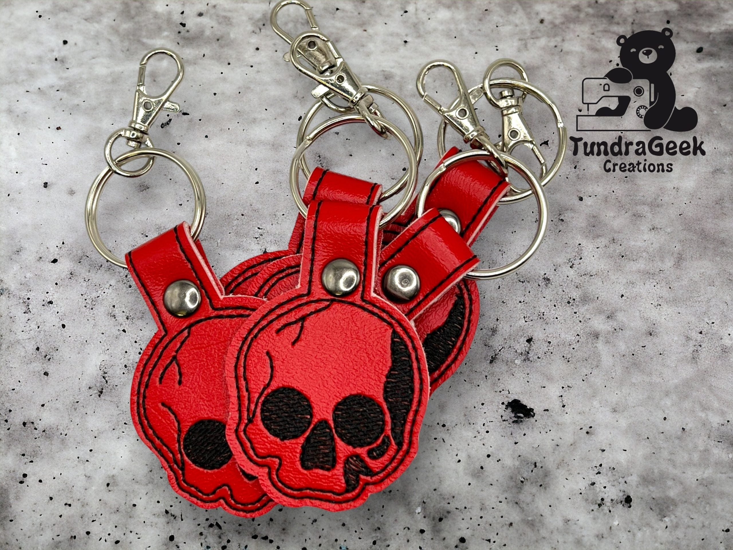Three red faux leather skull keychains with metal rings and clips are arranged on a textured surface. A TundraGeek Creations logo appears in the upper right corner.