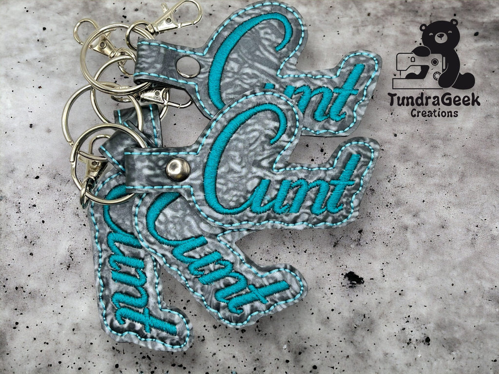 Three gray and teal keychains with the word Cunt in cursive, displayed on a gray surface with the TundraGeek Creations logo in the top right corner.