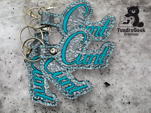 Three gray and teal keychains with the word Cunt in cursive, displayed on a gray surface with the TundraGeek Creations logo in the top right corner.