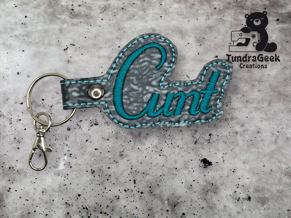 A keychain with the word Cunt embroidered in teal thread on gray fabric, featuring a metal key ring and clasp. TundraGeek Creations logo is in the top right corner.
