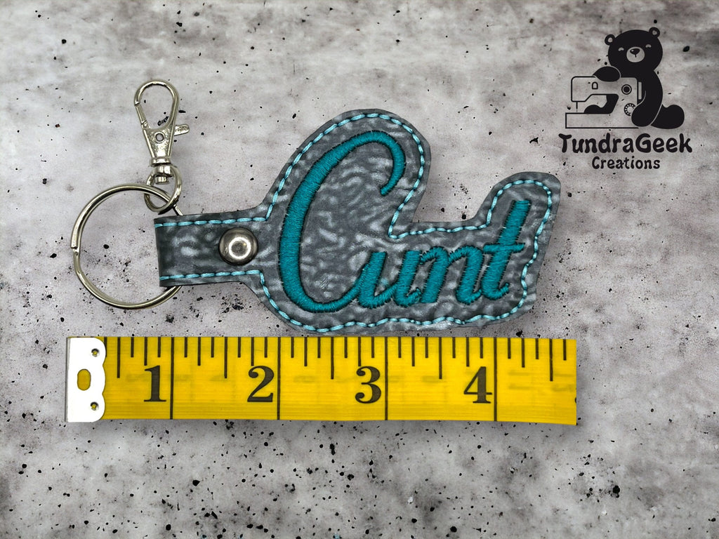 A gray and teal keychain with the word Cunt stitched on it is placed next to a yellow measuring tape, about four inches long. A logo reading TundraGeek Creations is in the top right corner.