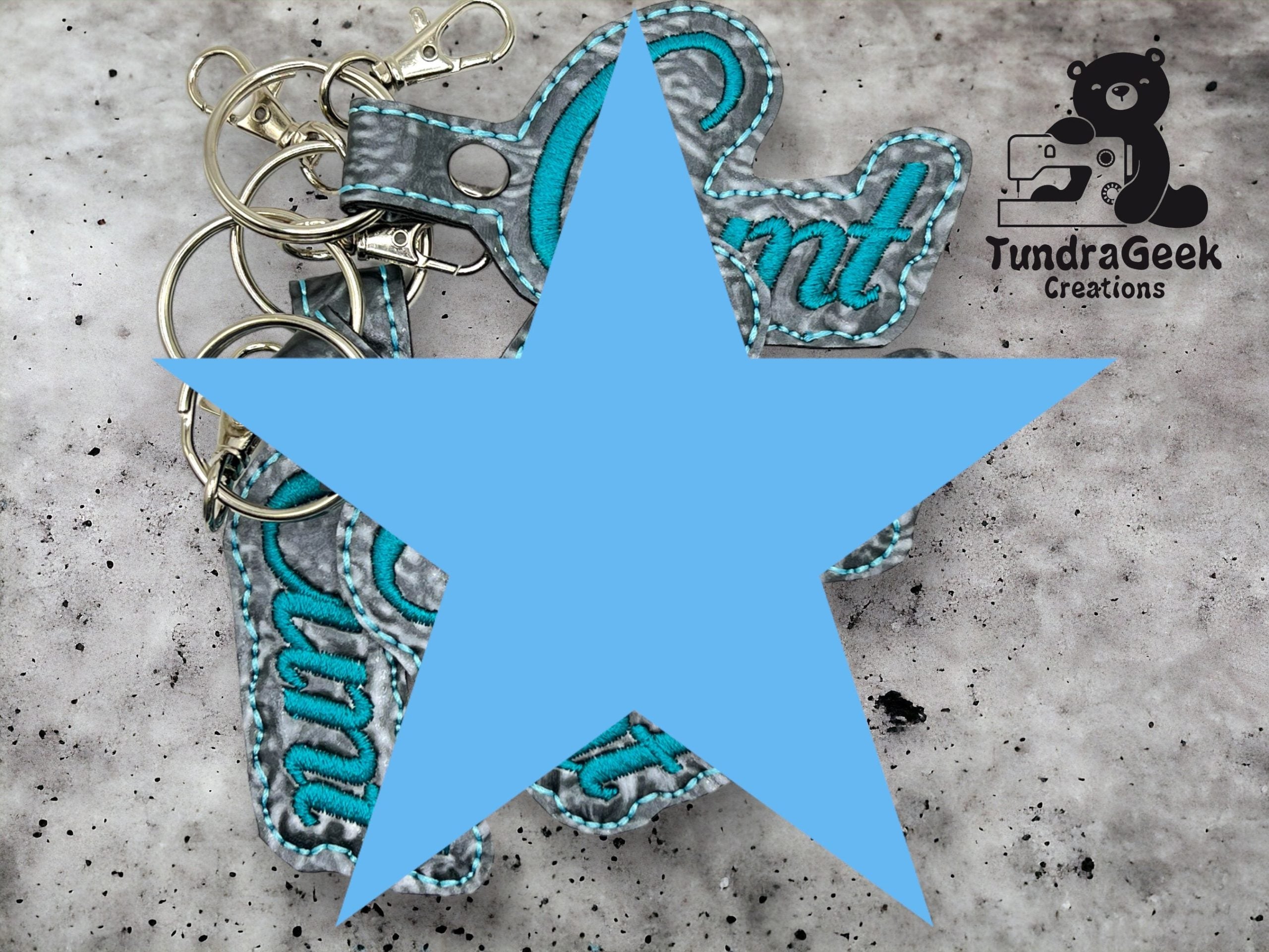Several keychains with embroidered text are arranged on a gray surface. The TundraGeek Creations logo appears in the top right corner. A large blue star covers part of the image.