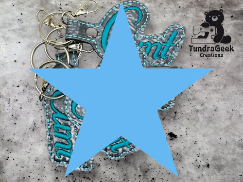 Several keychains with embroidered text are arranged on a gray surface. The TundraGeek Creations logo appears in the top right corner. A large blue star covers part of the image.