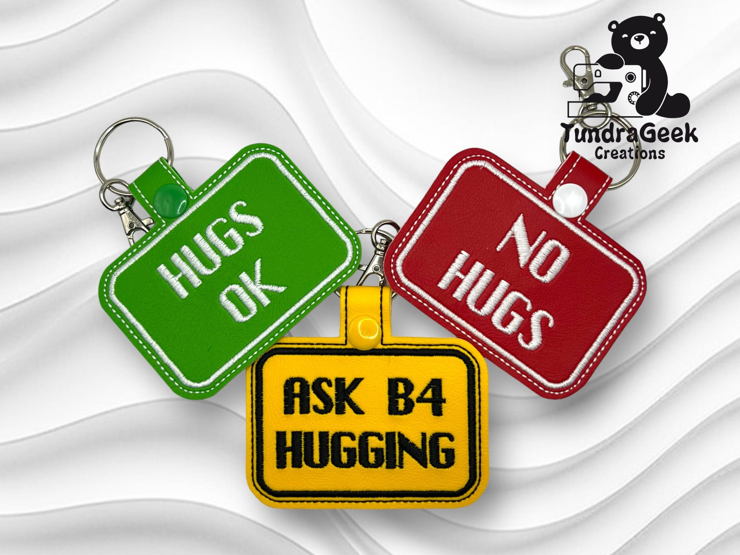 Three rectangular keychains with messages: HUGS OK (green), NO HUGS (red), and ASK B4 HUGGING (yellow) on a white textured background. Tundra Geek Creations logo in corner.