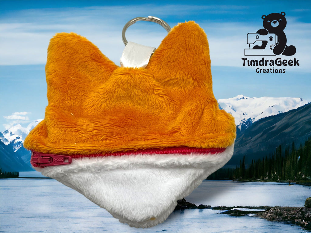 A small, plush pouch shaped like animal ears with an orange top, white bottom, and a red zipper, hanging from a keyring in front of a lake and mountain background.