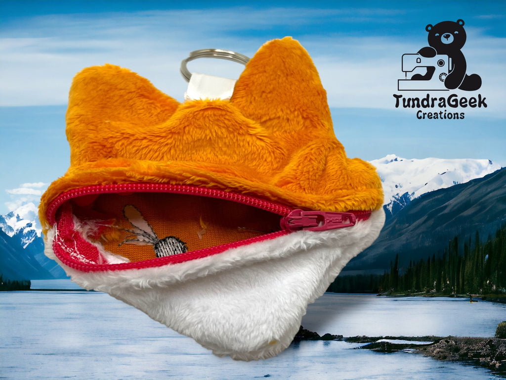 A small, plush fox-shaped keychain coin purse with a red zipper is displayed in front of a scenic mountain lake background with the TundraGeek Creations logo in the top right corner.