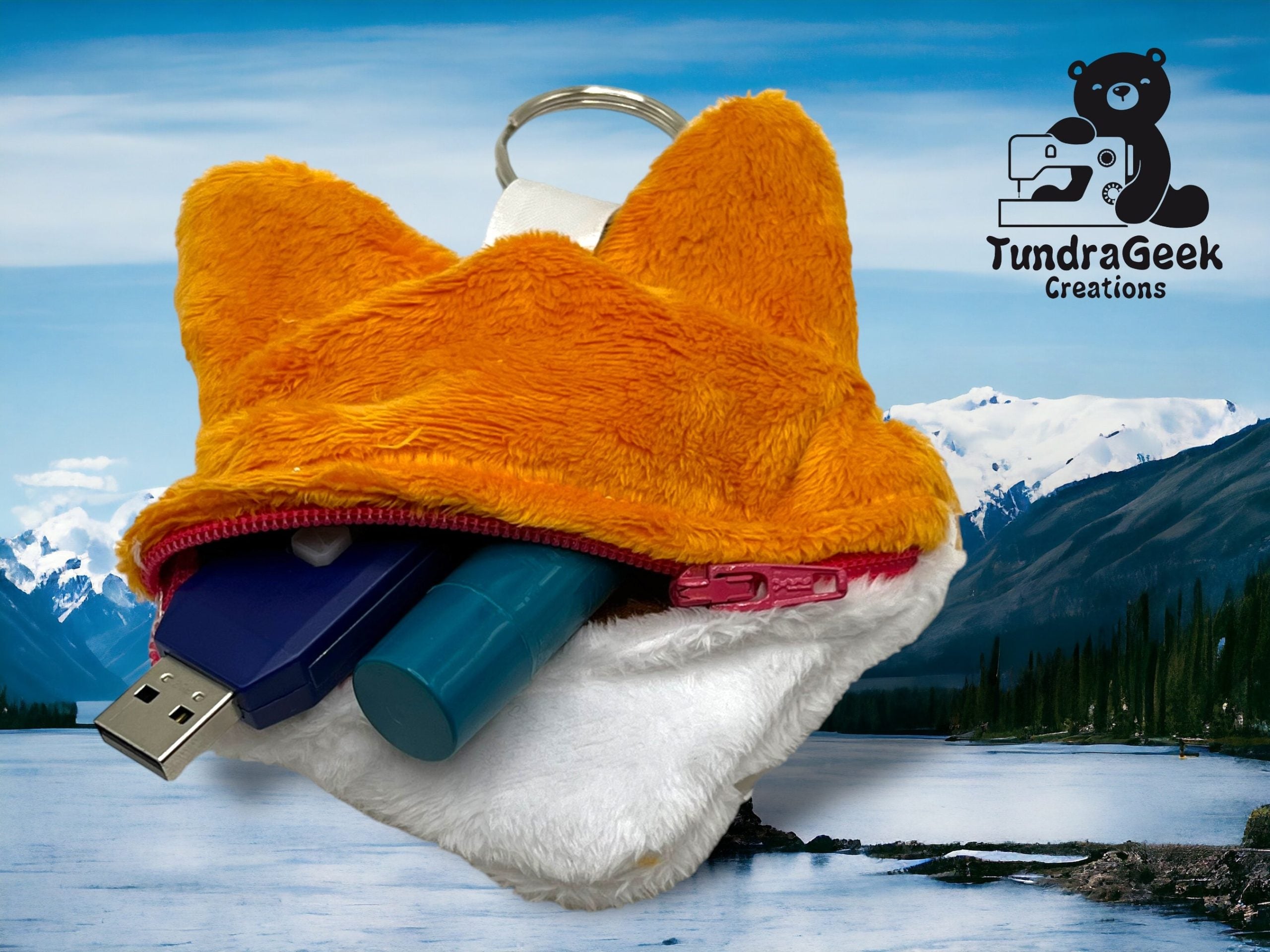 A small, fox-shaped plush keychain pouch with a zipper is open, showing a USB drive and lip balm inside. It sits in front of a lake and mountain landscape with a TundraGeek logo.