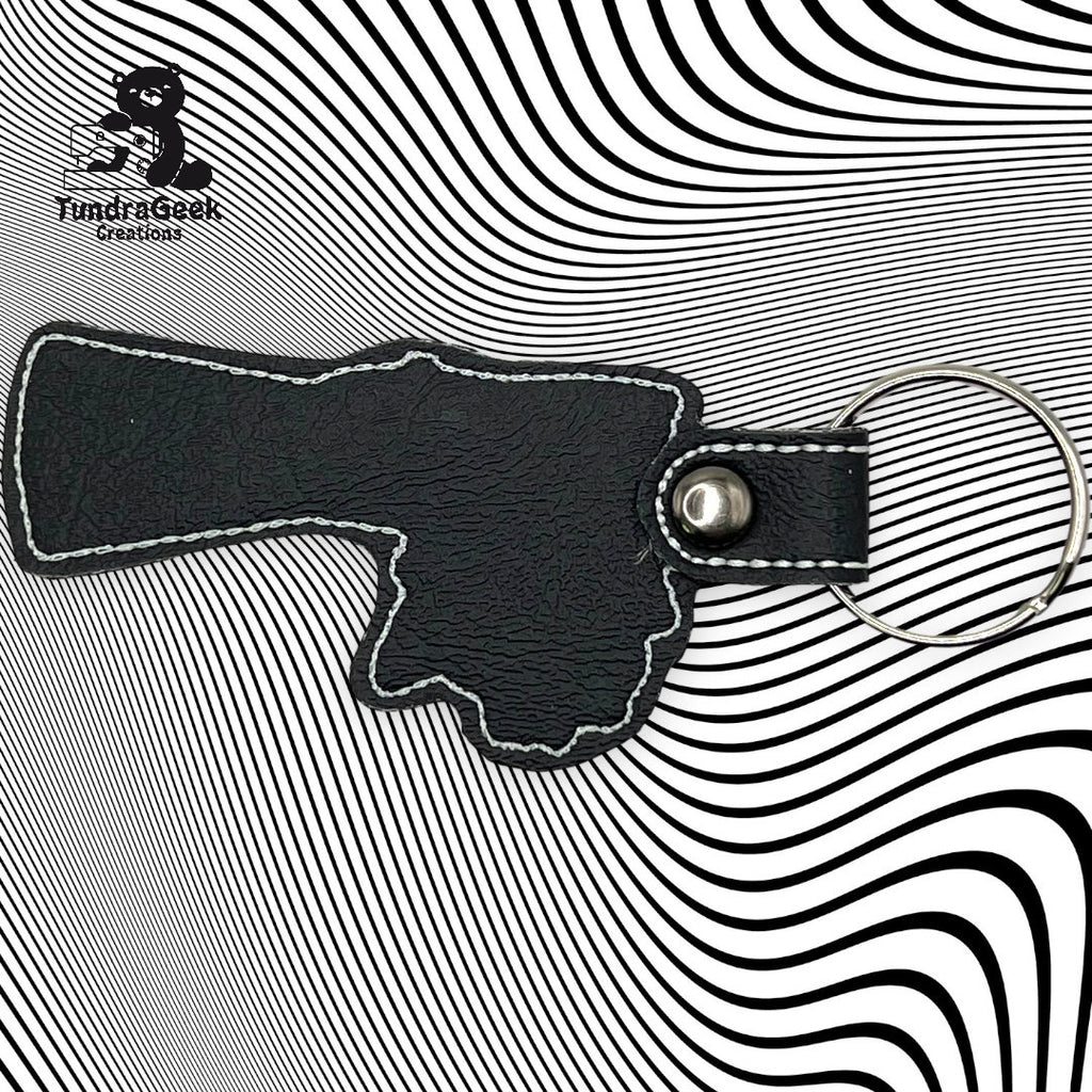 A black leather keychain shaped like a gun is placed on a black-and-white wavy striped background. The FundriGeek Creations logo appears in the upper left corner.