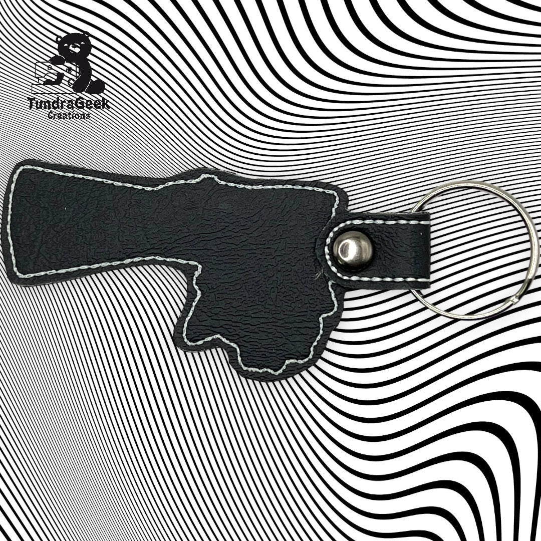 A black leather keychain shaped like a gun is placed on a black-and-white wavy striped background. The FundriGeek Creations logo appears in the upper left corner.
