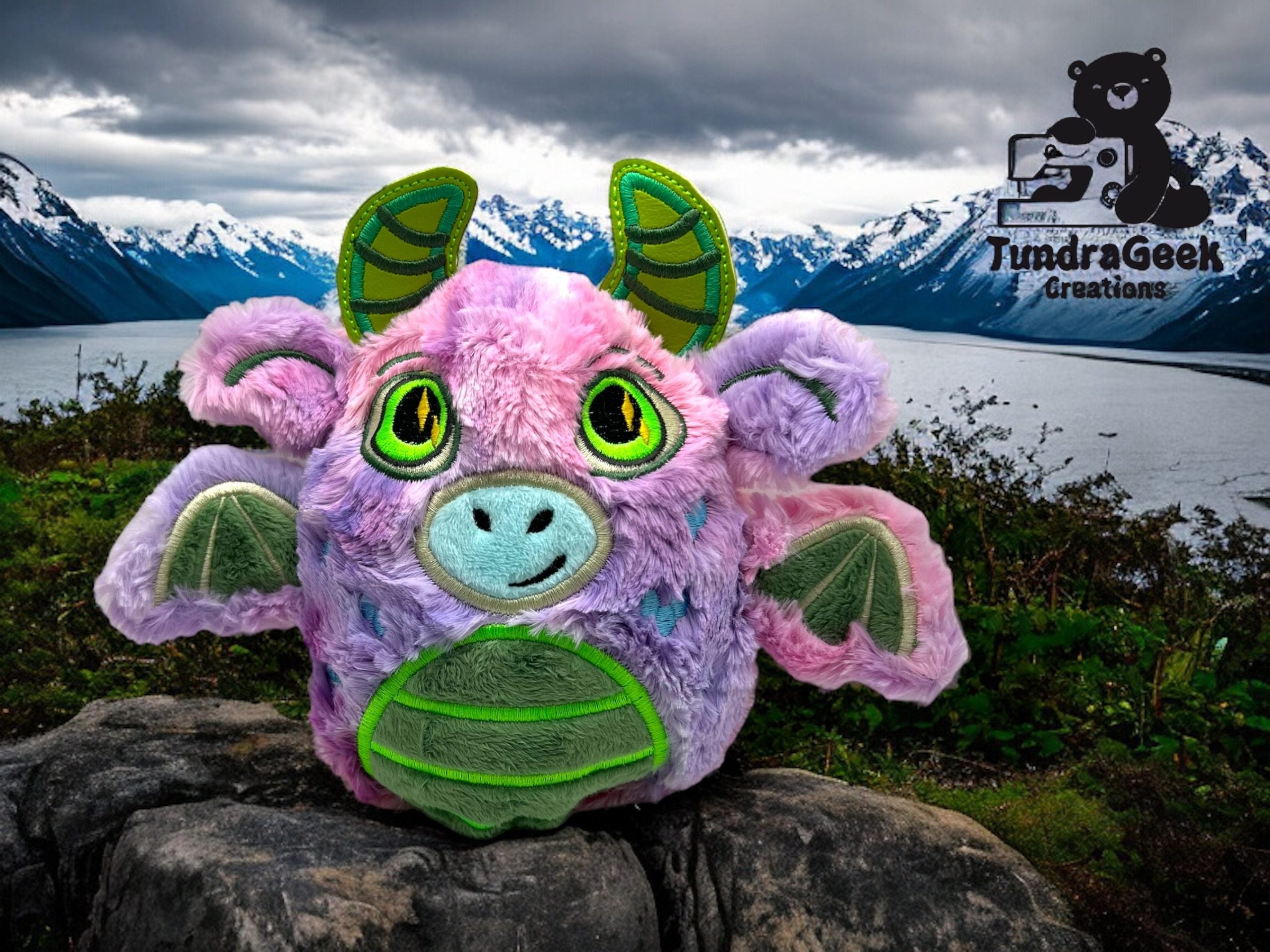A plush dragon toy with pink, green, and purple colors sits on a rock with mountains, lake, and cloudy sky in the background. TundraGeek Creations logo appears in the corner.