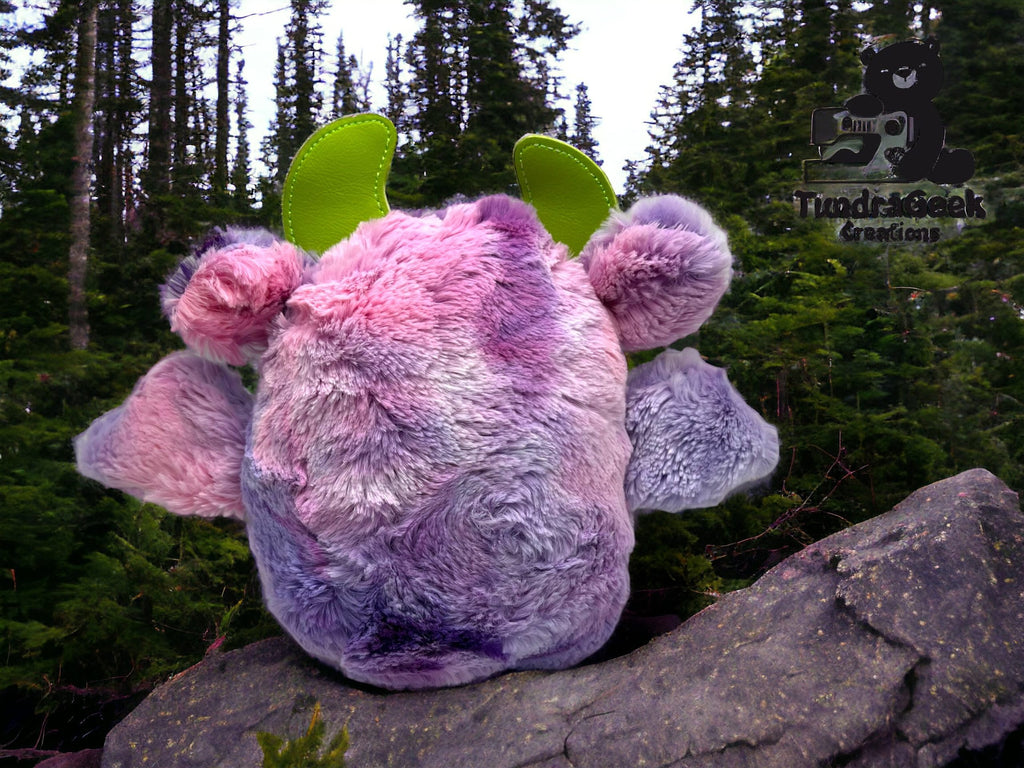A round, pastel-colored plush toy with green horns sits on a rock in front of evergreen trees under a cloudy sky.