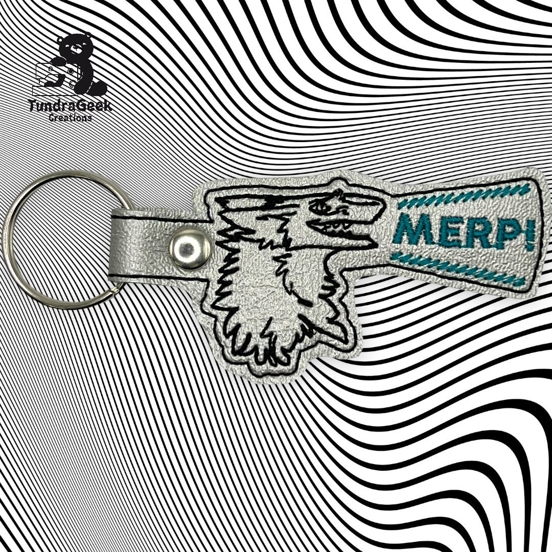 Keychain with a stylized wolf head and the word MERP! in blue, placed on a black and white wavy line background. Logo reading Tundra Geek Creations in top left corner.