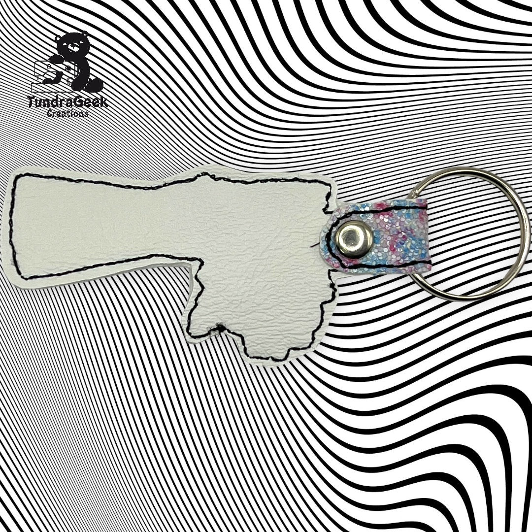 A keychain in the shape of Oklahoma with a metal ring and snap, placed on a black and white wavy patterned background. FundrisGeek Creations logo is in the top left corner.