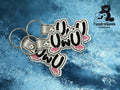 Two embroidered keychains with owo text and bunny ear designs are placed on a blue marbled surface, next to the TundraGeek Creations logo.