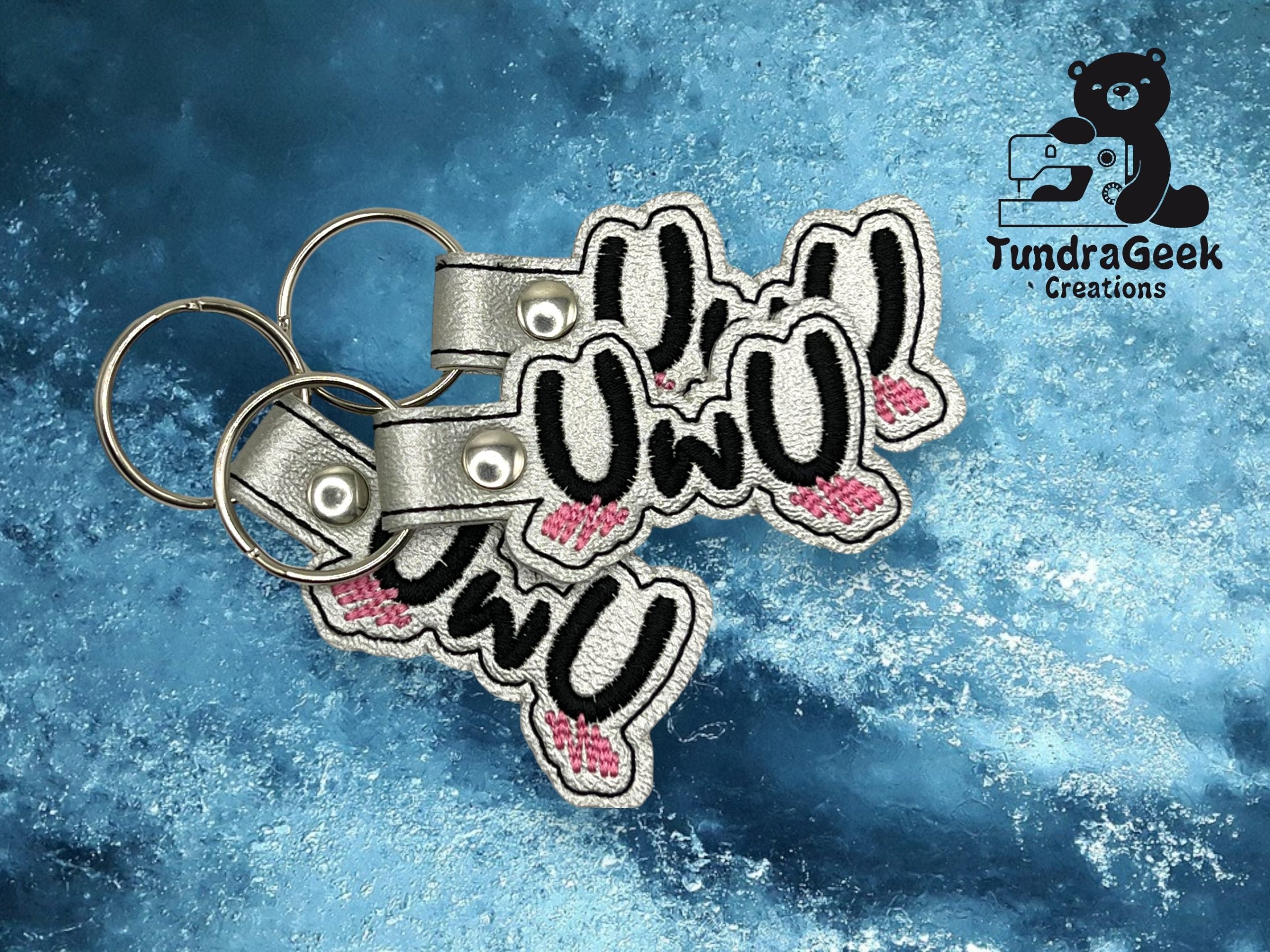 Two embroidered keychains with owo text and bunny ear designs are placed on a blue marbled surface, next to the TundraGeek Creations logo.