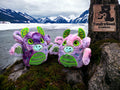 Two plush dragon toys sit on mossy ground by a partially frozen lake with snow-capped mountains in the background; a TundraGeek Creations sign is attached to a tree.