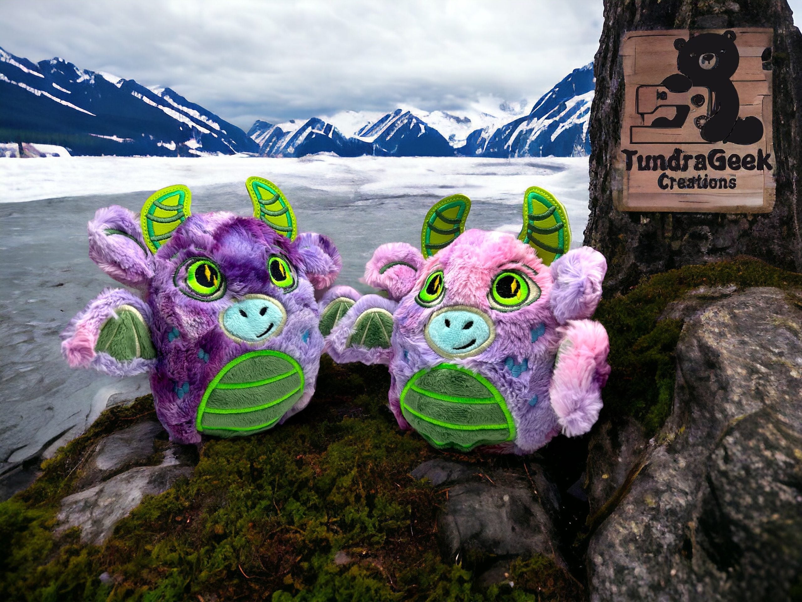 Two plush dragon toys sit on mossy ground by a partially frozen lake with snow-capped mountains in the background; a TundraGeek Creations sign is attached to a tree.