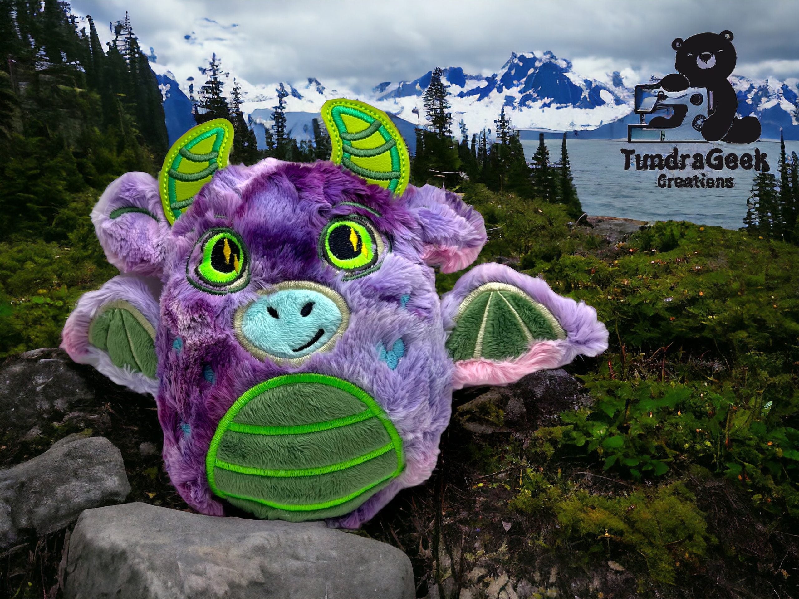 A plush dragon toy with purple, green, and blue colors sits on rocks in a mountainous landscape with a lake, trees, and snow-capped peaks. TundraGeek Creations logo is in the corner.