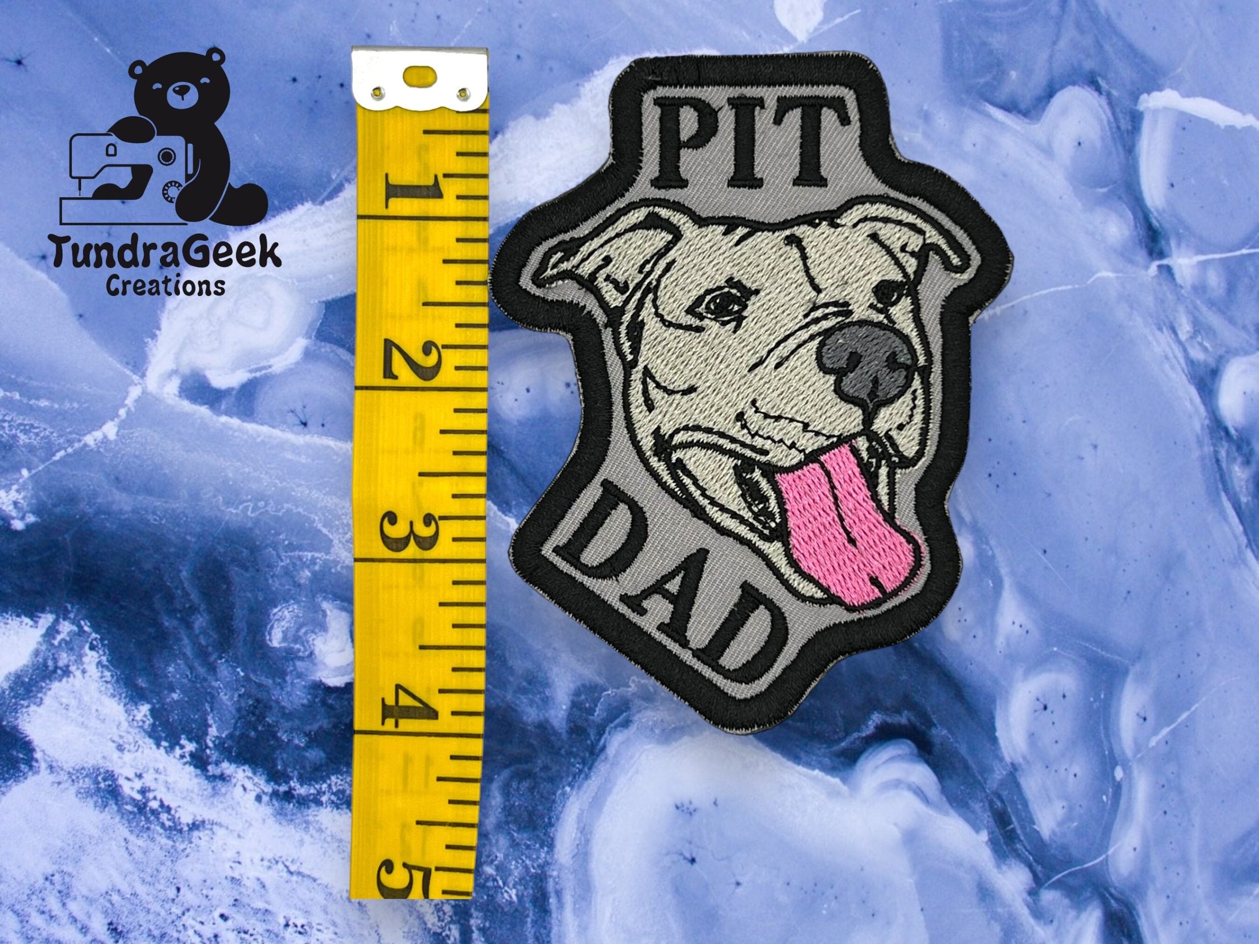 An embroidered patch with a pit bull face and the text PIT DAD next to a yellow tape measure showing roughly 3 inches, on a marbled blue background.