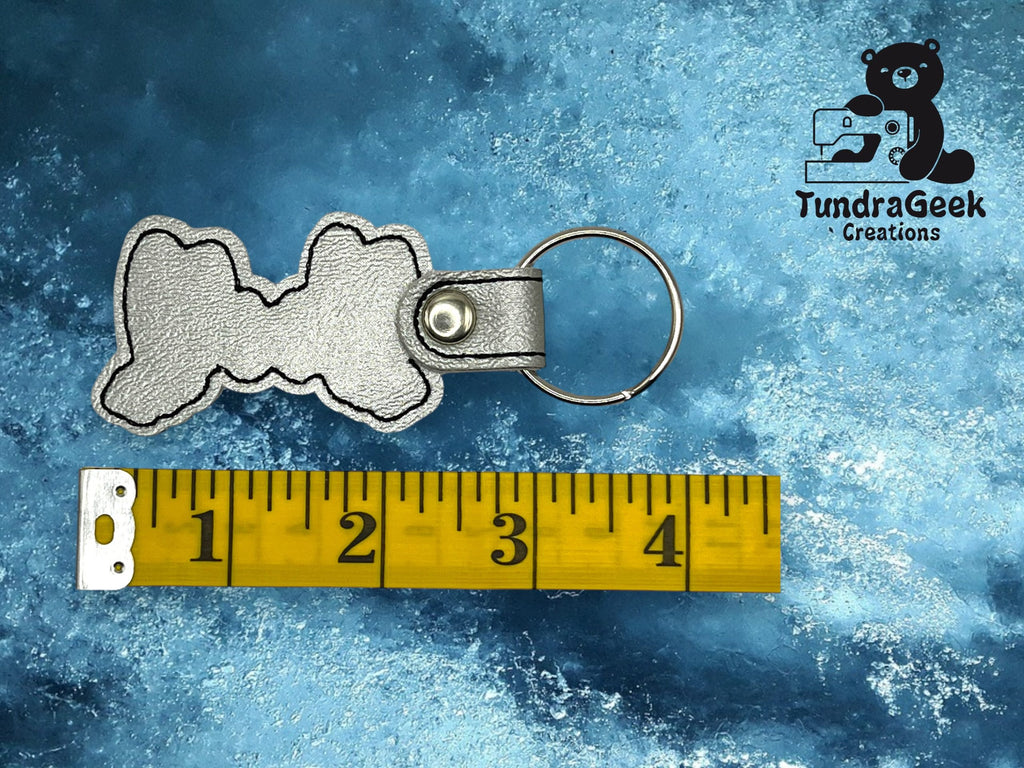 A silver dog bone-shaped keychain is next to a measuring tape showing about 3 inches, with the TundraGeek Creations logo in the top right corner.