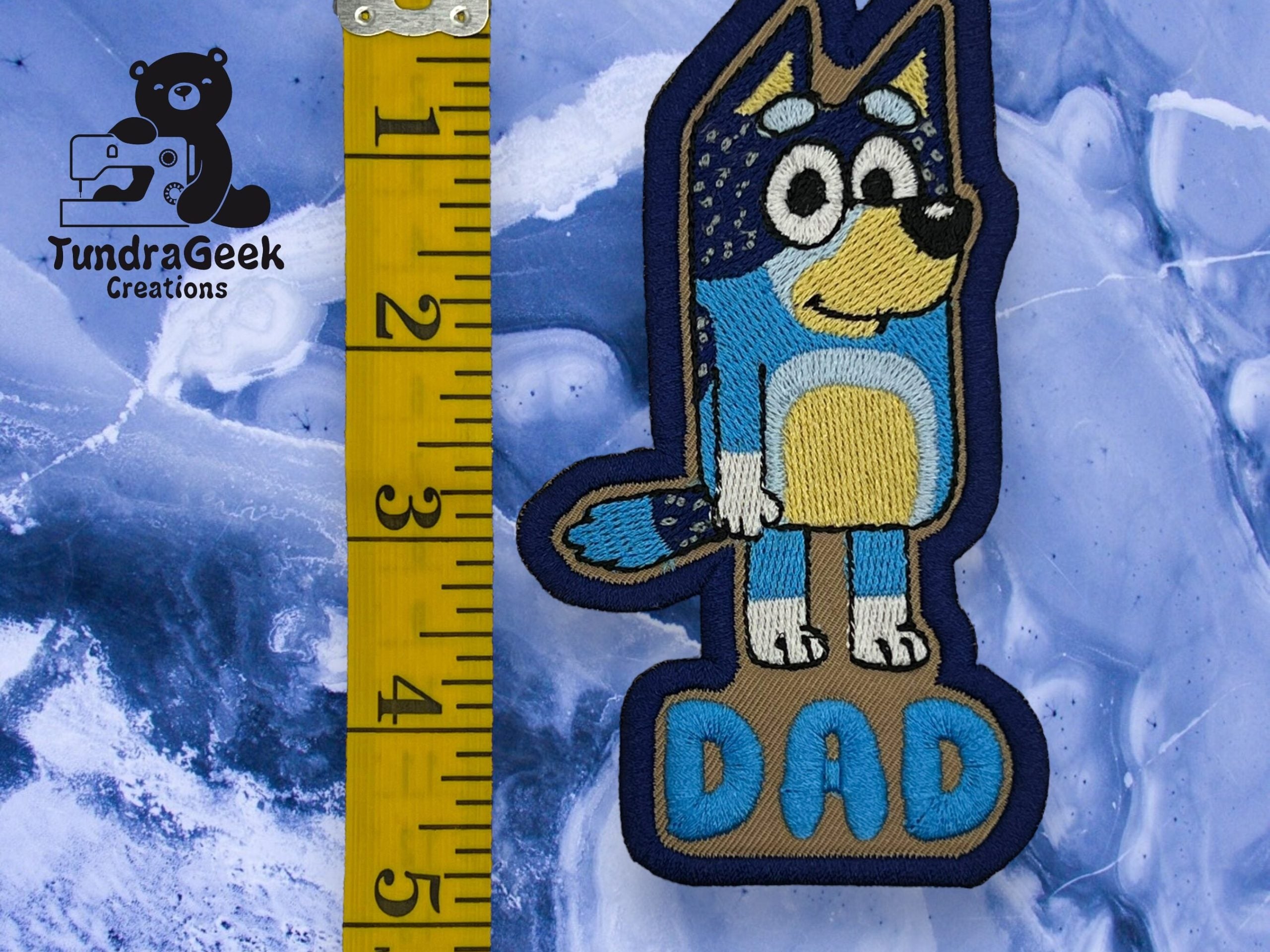 Embroidered patch of a cartoon blue dog labeled DAD next to a yellow measuring tape on a blue marbled background, with TundraGeek Creations logo in the corner.