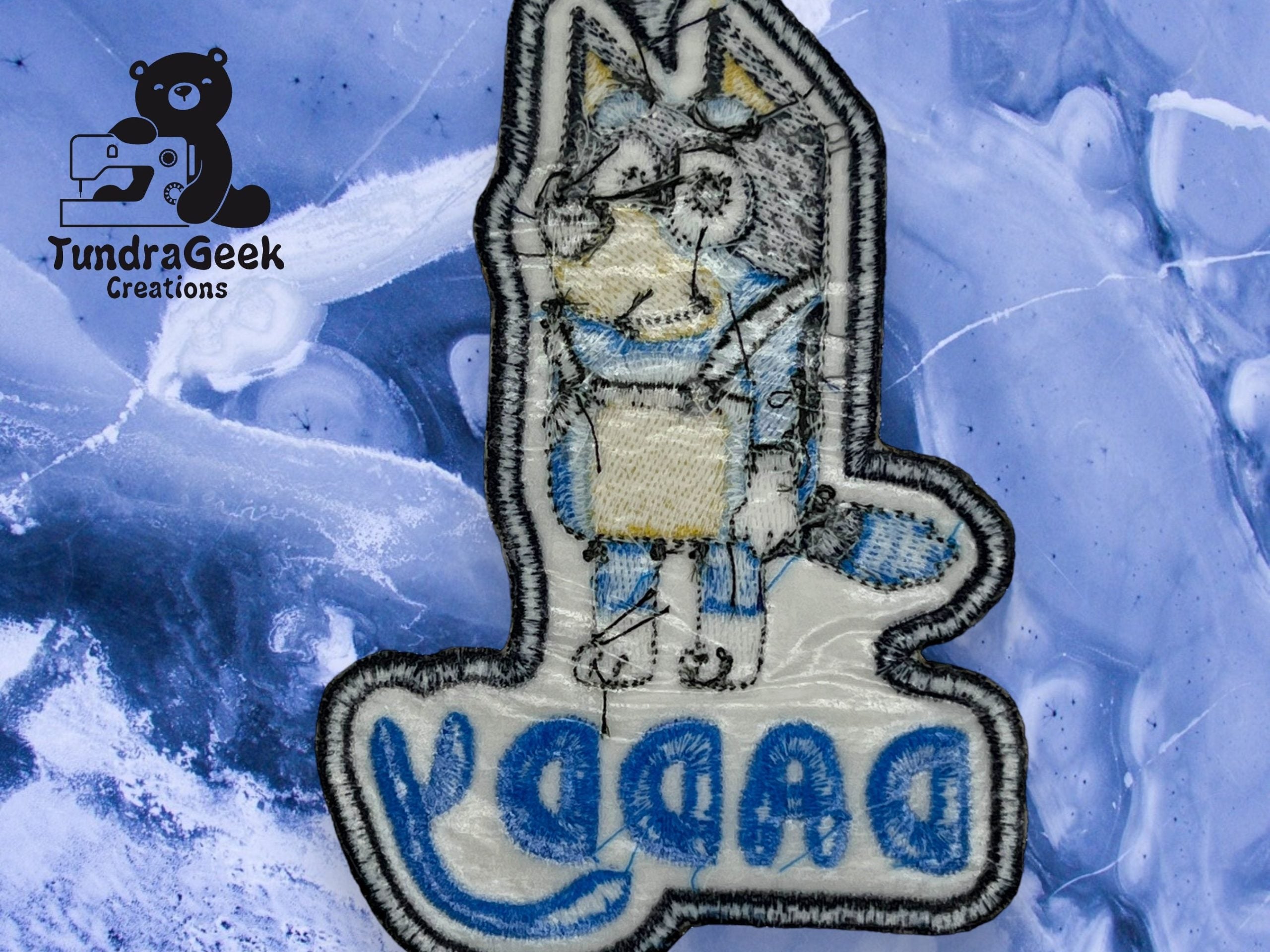 Embroidered patch of a cartoon animal with the word DADDY on it, placed on a blue marbled surface. TundraGeek Creations logo appears in the top left corner.