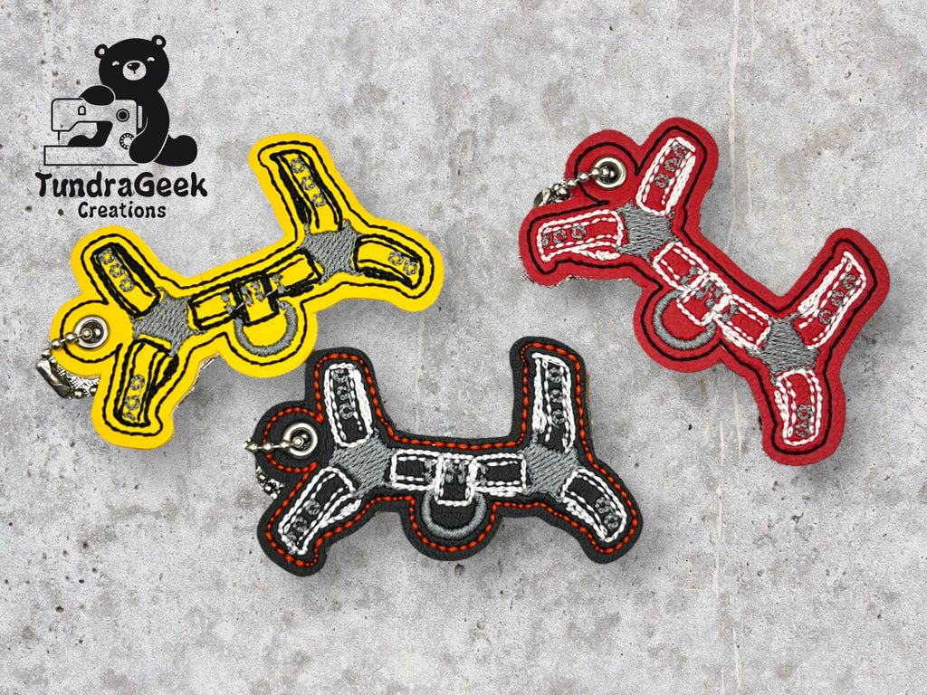 Three keychains shaped like dog harnesses in yellow, red, and black are displayed on a gray stone surface. A TundraGeek Creations logo is in the top left corner.