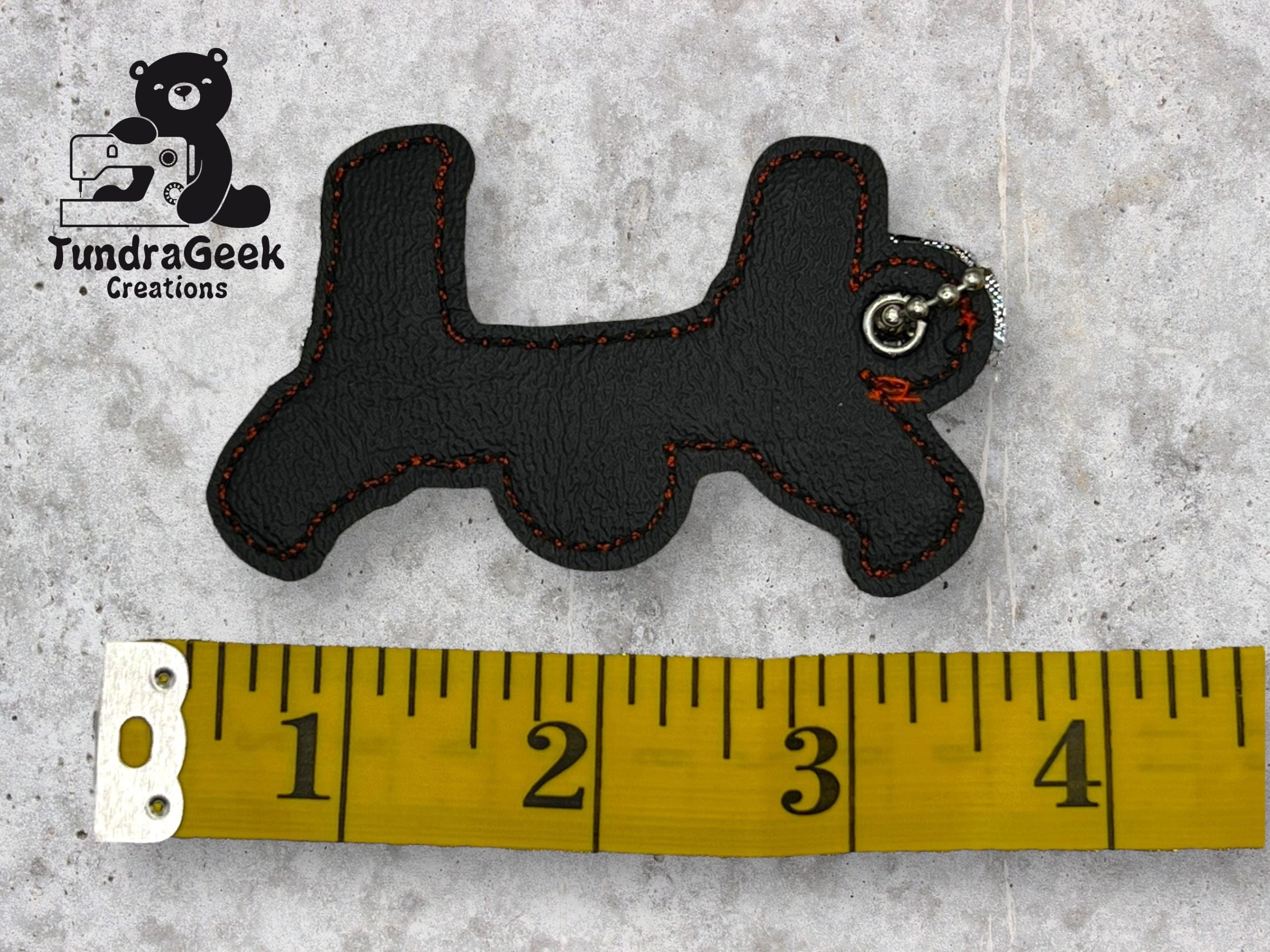 A keychain shaped like a dog, made of black material with red stitching, lies next to a measuring tape showing a length of about 3 inches. The TundraGeek Creations logo is in the top left.