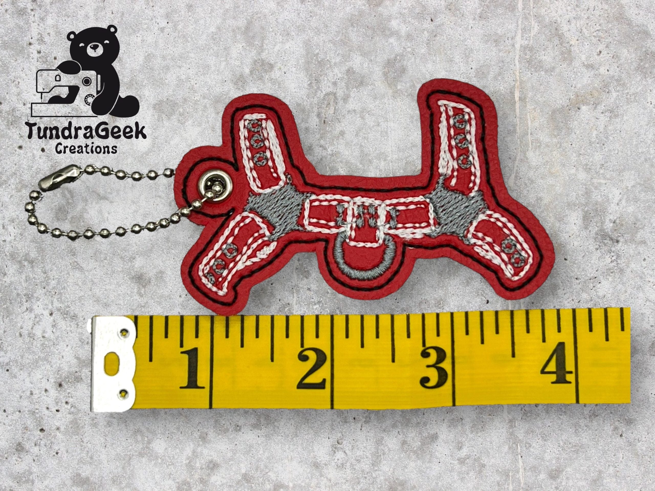 Embroidered red and gray keychain shaped like a dog harness next to a yellow measuring tape showing a length of about 3 inches. TundraGeek Creations logo in the corner.