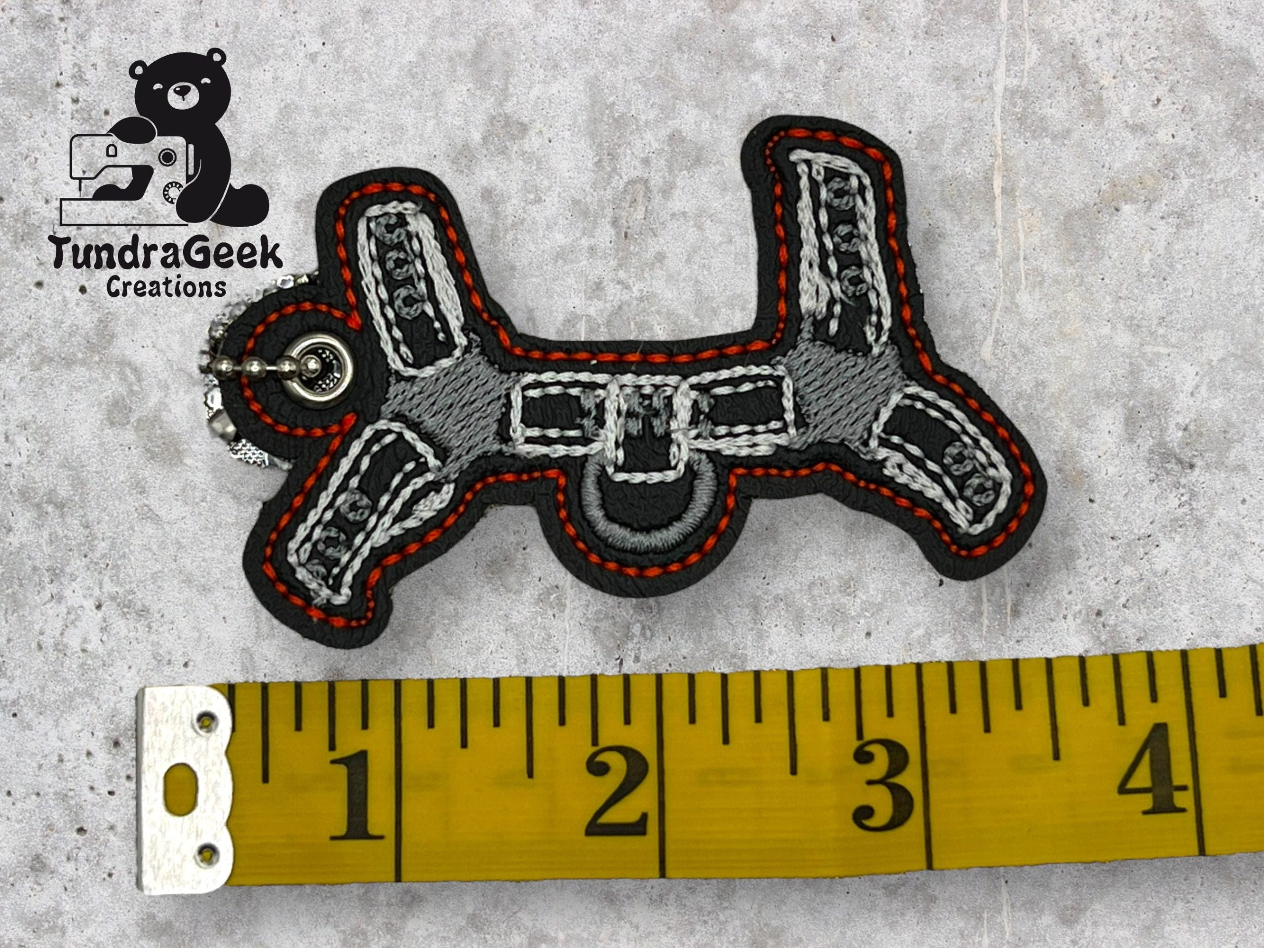 Embroidered keychain in the shape of a dog with a metal ring, placed above a yellow measuring tape showing about 3 inches. TundraGeek Creations logo is in the top left corner.
