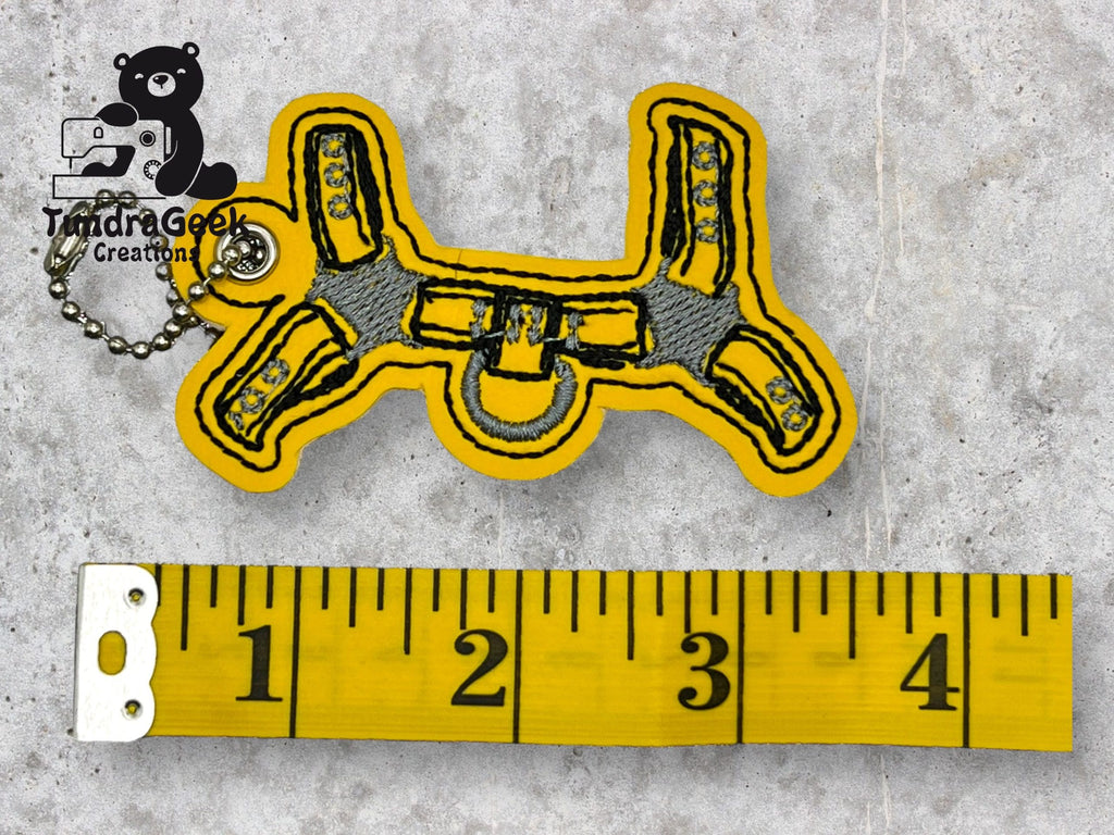 Keychain shaped like a climbing harness with yellow and gray stitching next to a measuring tape showing a length of about 3 inches. TundraGeek Creations logo in the corner.