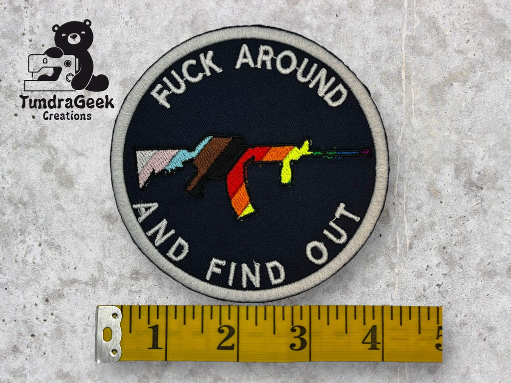 A circular patch with an illustrated rifle in rainbow colors and the text FUCK AROUND AND FIND OUT, next to a yellow measuring tape and a TundraGeek Creations logo.