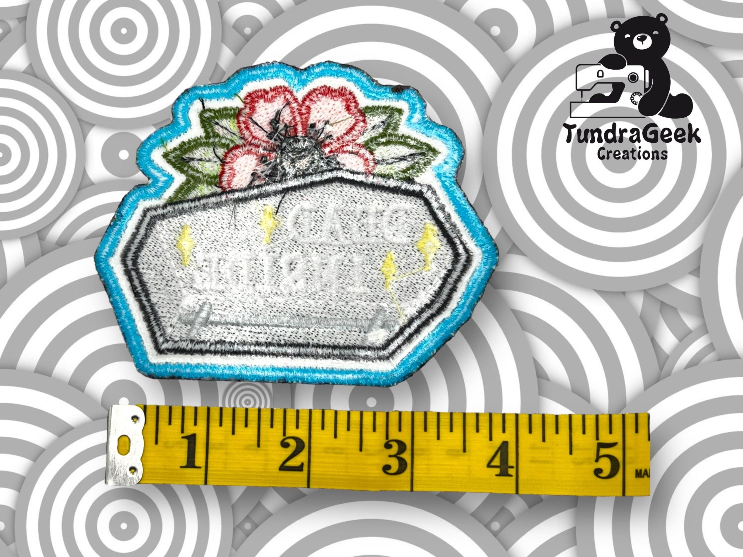 An embroidered patch shaped like a coffin with flowers on top is shown next to a measuring tape; background features concentric circles and a bear logo reading TundraGeek Creations.