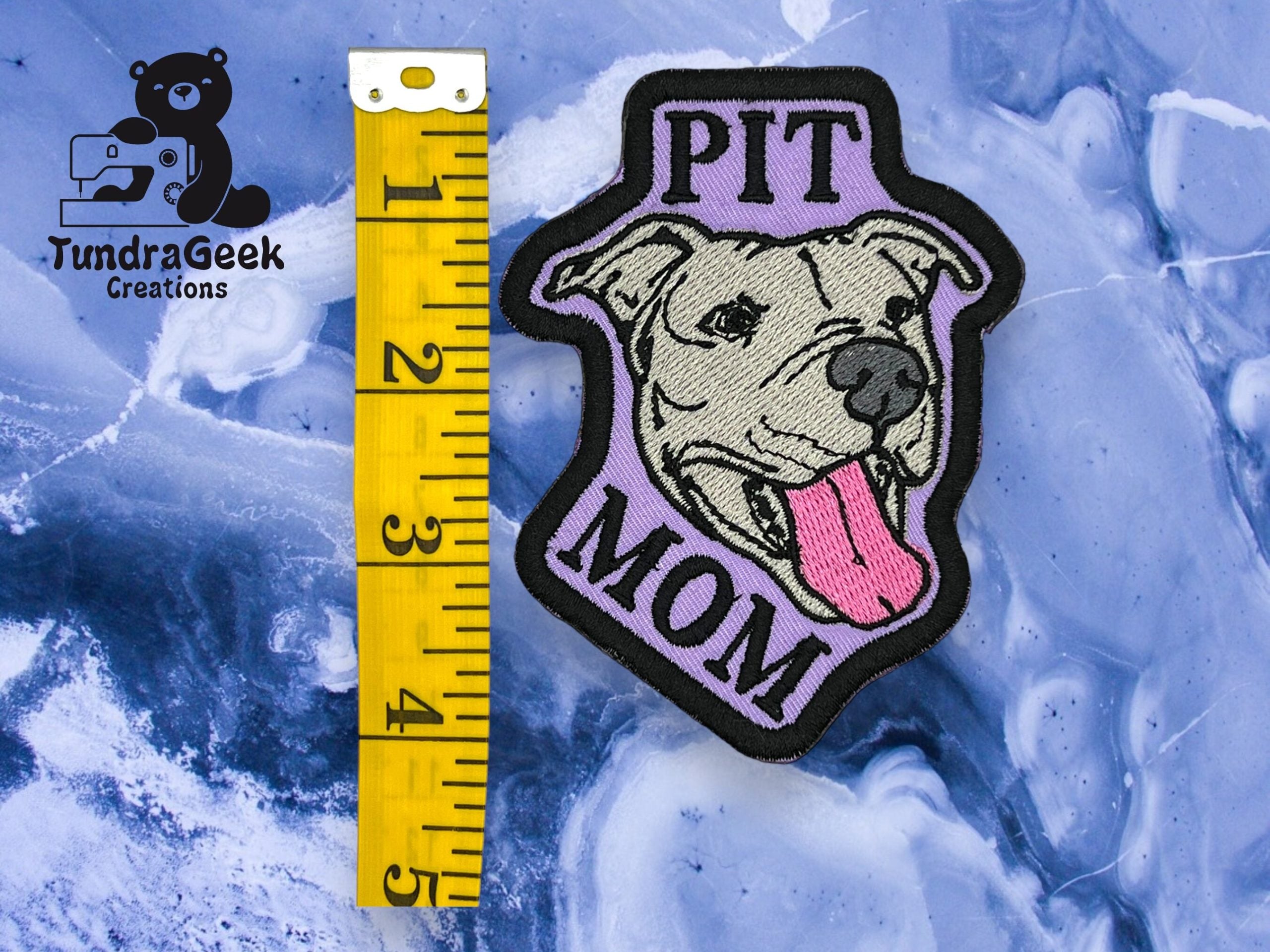 A patch reading PIT MOM with a cartoon pit bulls face next to a yellow measuring tape on a blue marbled surface. TundraGeek Creations logo is in the top left corner.