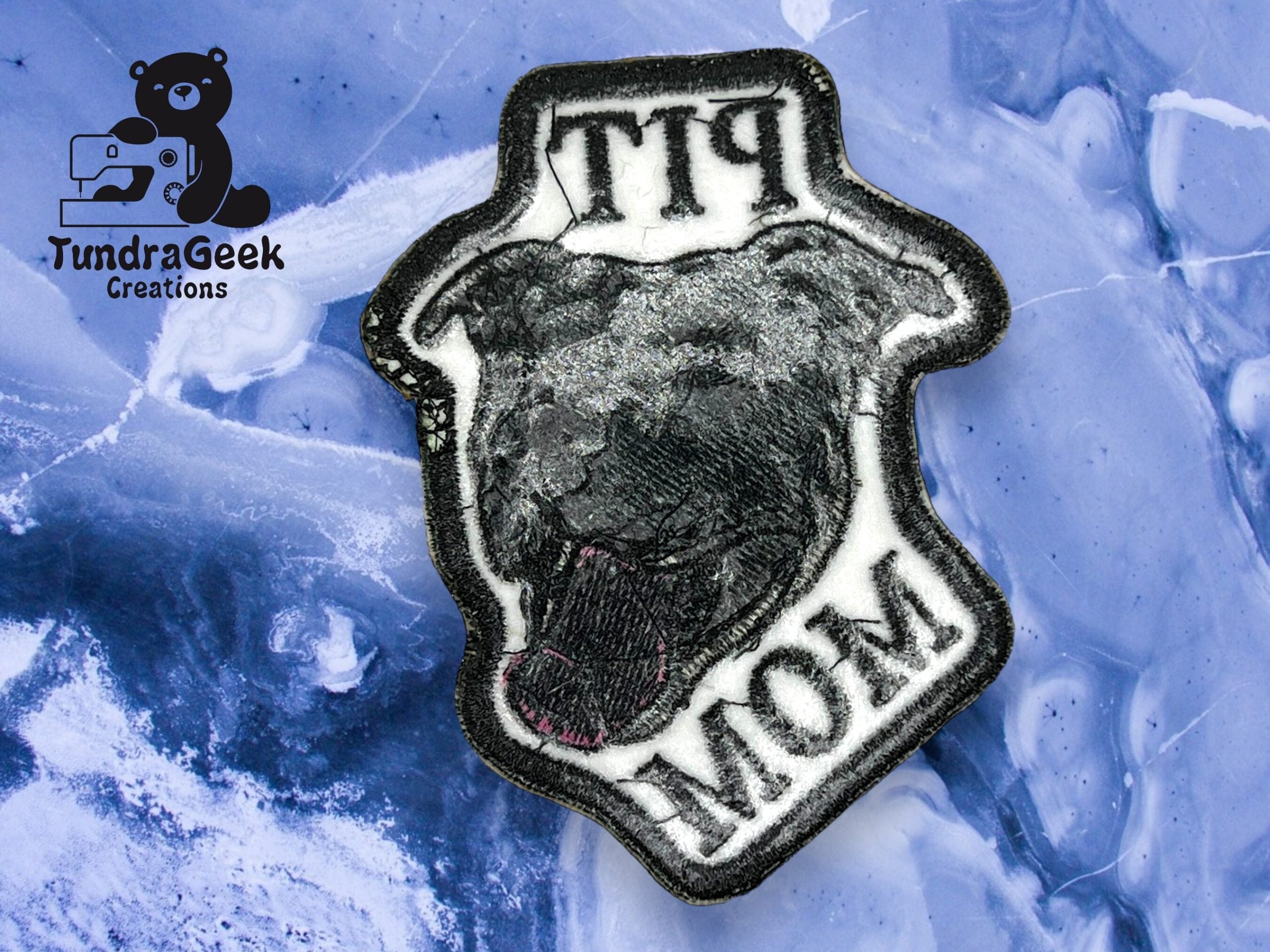 An embroidered patch showing a black dogs head with PIT MOM text, placed on a blue and white marbled surface. A TundraGeek Creations logo is in the top left corner.