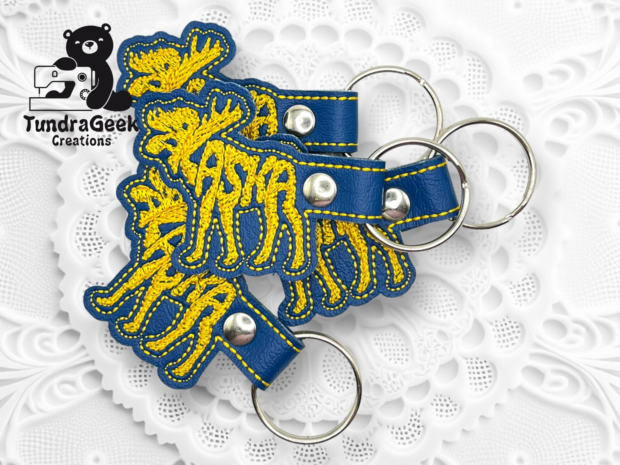 Several blue and yellow embroidered moose-shaped keychains with metal rings are arranged on a white patterned surface. TundraGeek Creations logo appears in the upper left corner.