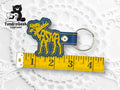 A blue and yellow embroidered moose-shaped keychain is placed next to a measuring tape showing a length of about 4 inches. The TundraGeek Creations logo appears in the upper left corner.