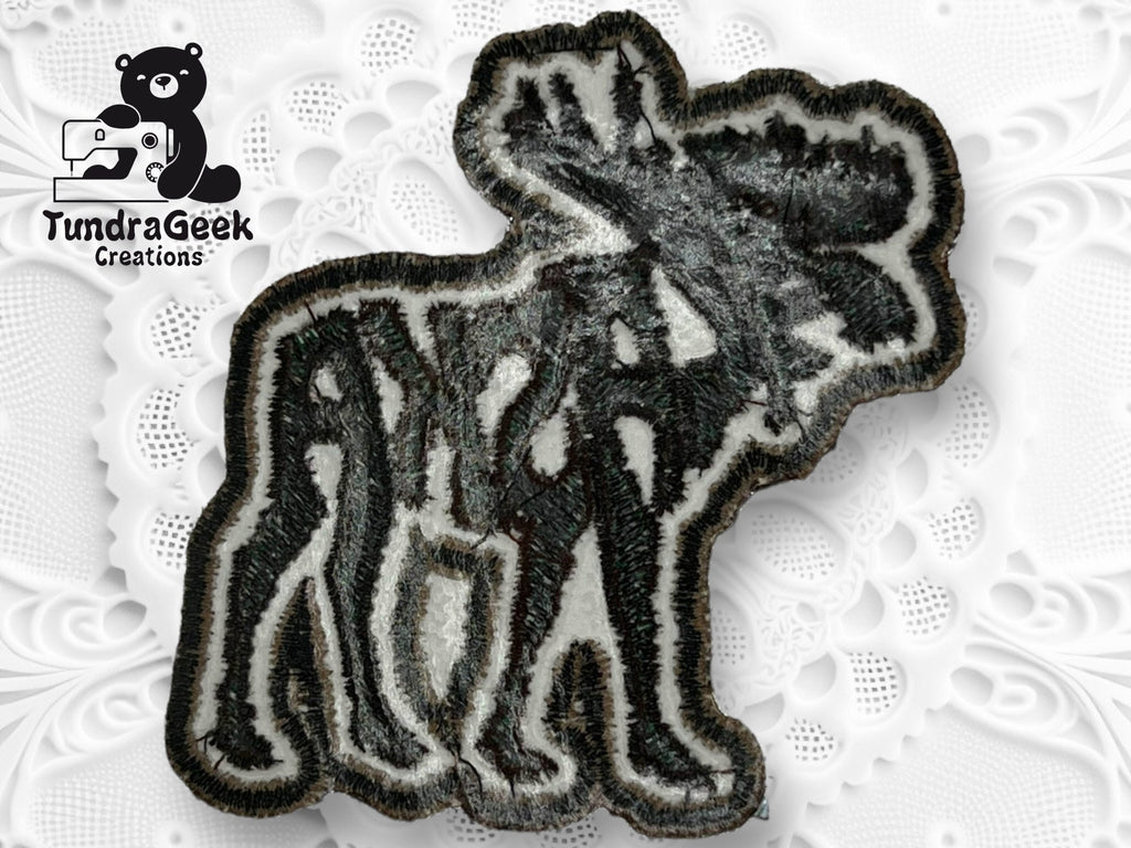 An embroidered patch shaped like a moose with the word Alaska integrated into its body, placed on a white textured surface with a TundraGeek Creations logo in the corner.