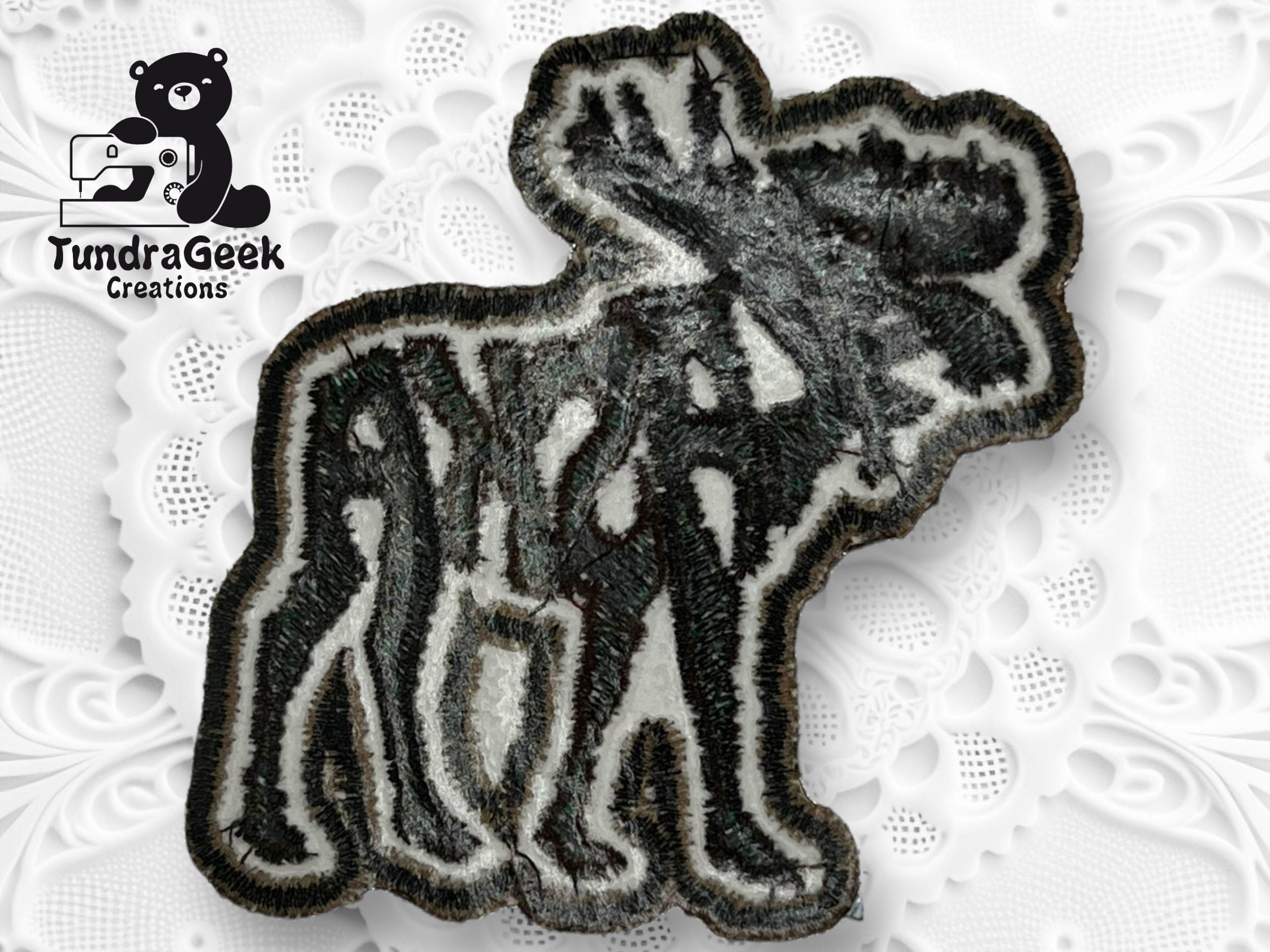 An embroidered patch shaped like a moose with the word Alaska integrated into its body, placed on a white textured surface with a TundraGeek Creations logo in the corner.