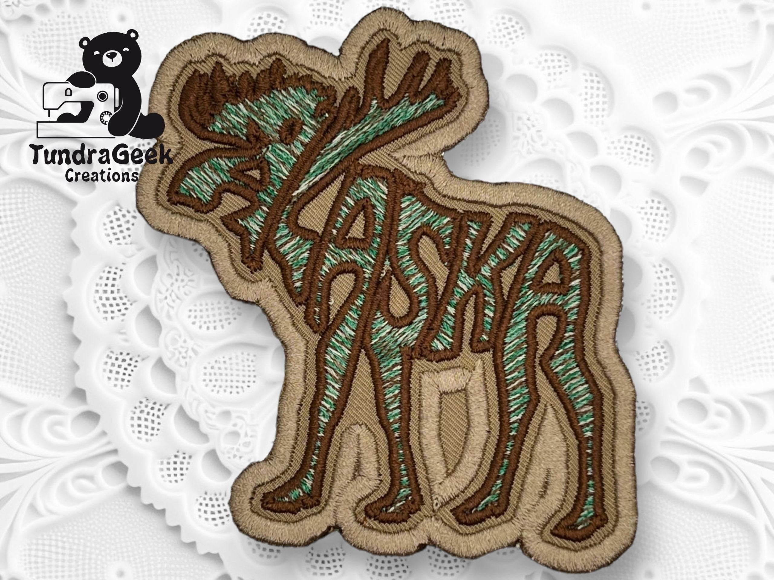 Embroidered patch of a moose with the word Alaska integrated into its body, placed on a decorative white background with the TundraGeek Creations logo in the corner.