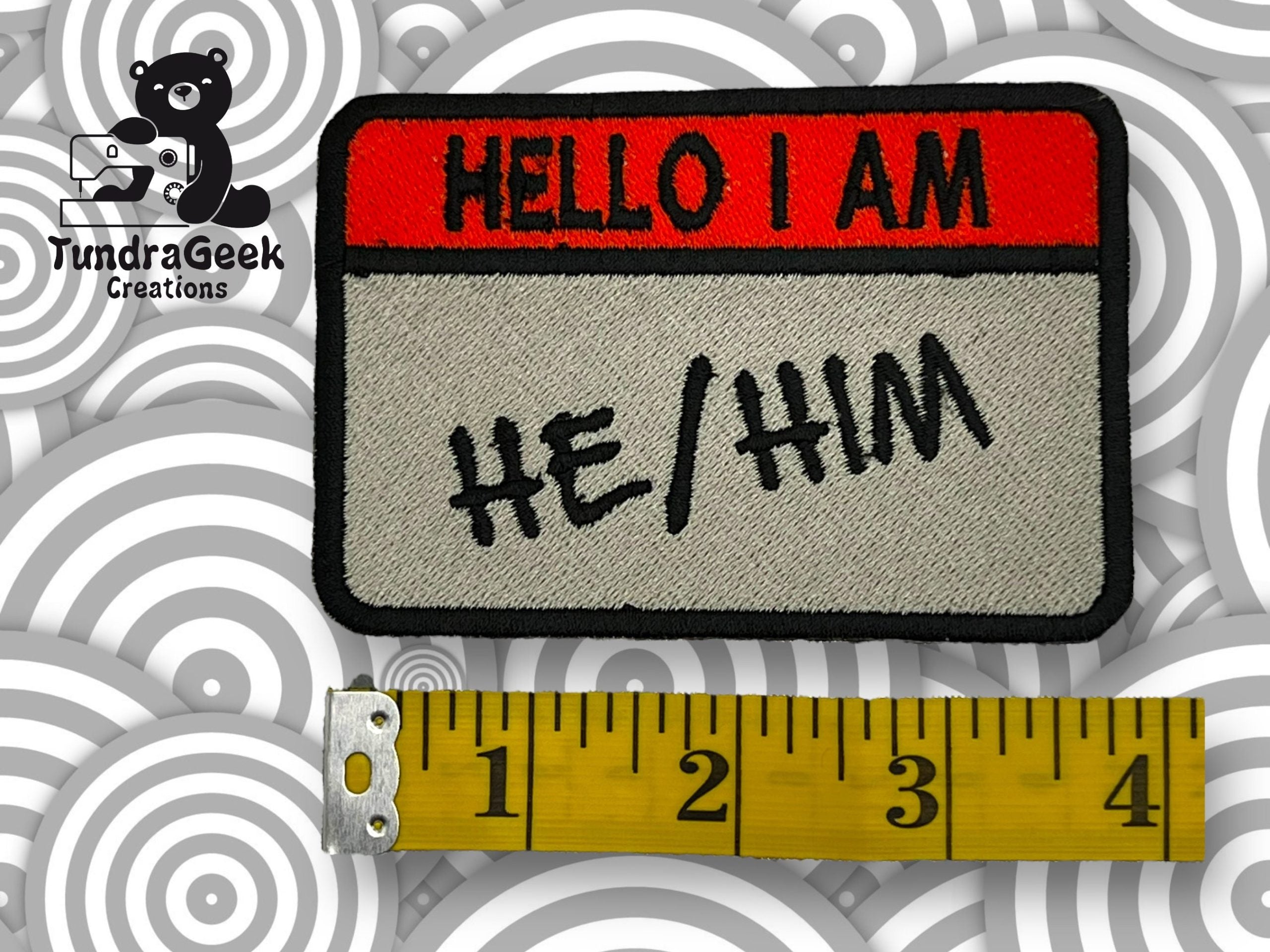 A rectangular patch reading HELLO I AM HE/HIM above a measuring tape showing about 4 inches, with a TundraGeek Creations logo in the top left corner.