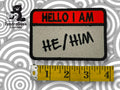 A Hello I am name tag patch with HE/HIM written on it, next to a yellow measuring tape showing a length of about four inches. The TundraGeek Creations logo is in the corner.